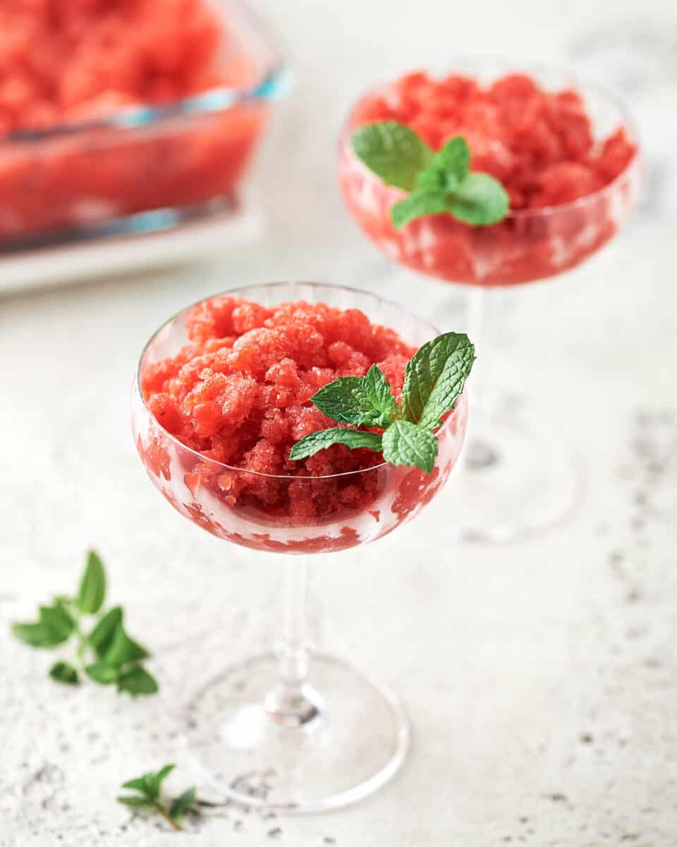 Side view of Strawberry Lemon Gin Granita in 2 coupe glasses with mint garnish with glass container in background