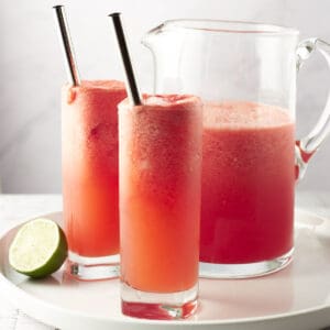 Side view of two glasses of watermelon coolers with pitcher on a white round plate