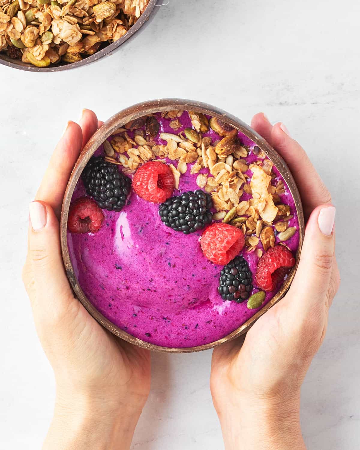 overhead view of hands holding dragonfruit smoothie bowl with granola, fresh berries