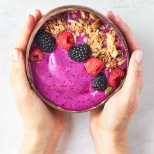 overhead view of hands holding dragonfruit smoothie bowl with granola, fresh berries with marble background