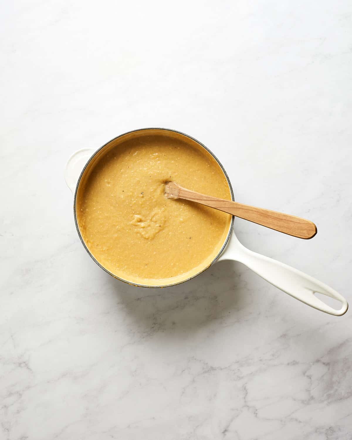 overhead view of cooking polenta in white saucepan with wooden spoon on marble background