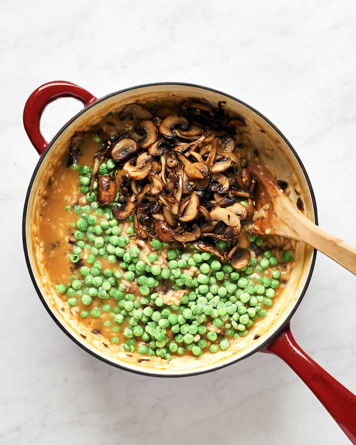 Cooked mushrooms and frozen peas added to risotto in saucepan on marble background
