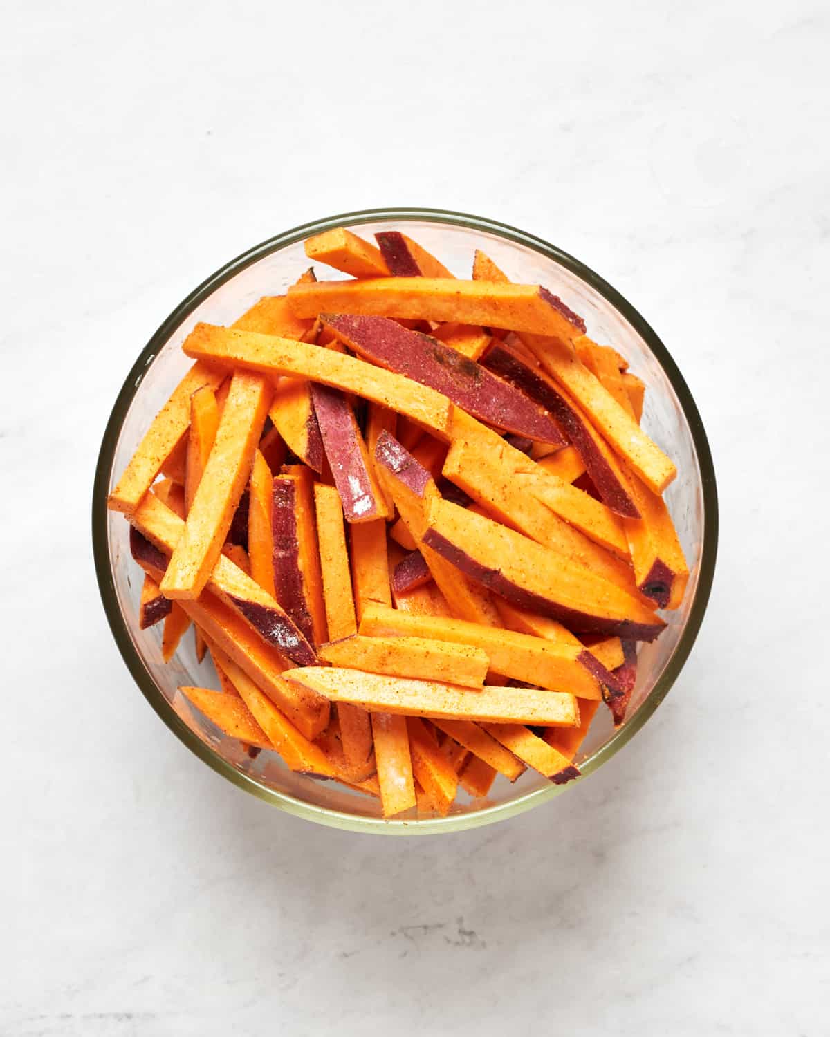 Freshly sliced sweet potato tossed with olive oil and spices in glass bowl