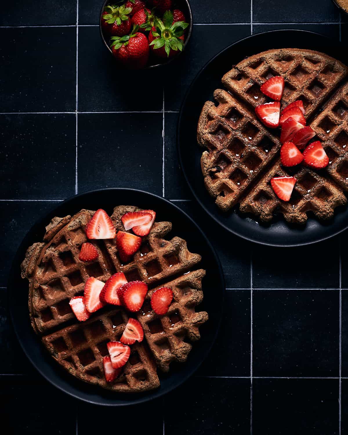 Overhead view of two plates of waffles with strawberries and maple syrup on top.