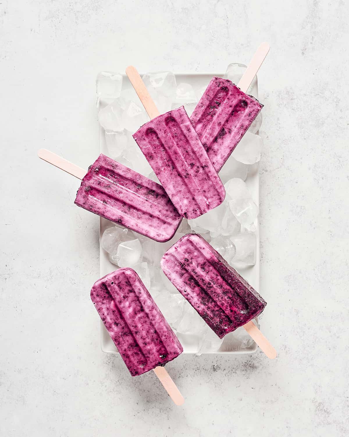 Overhead view of cherry coconut popsicles on ice on white tray on grey/white stone background.