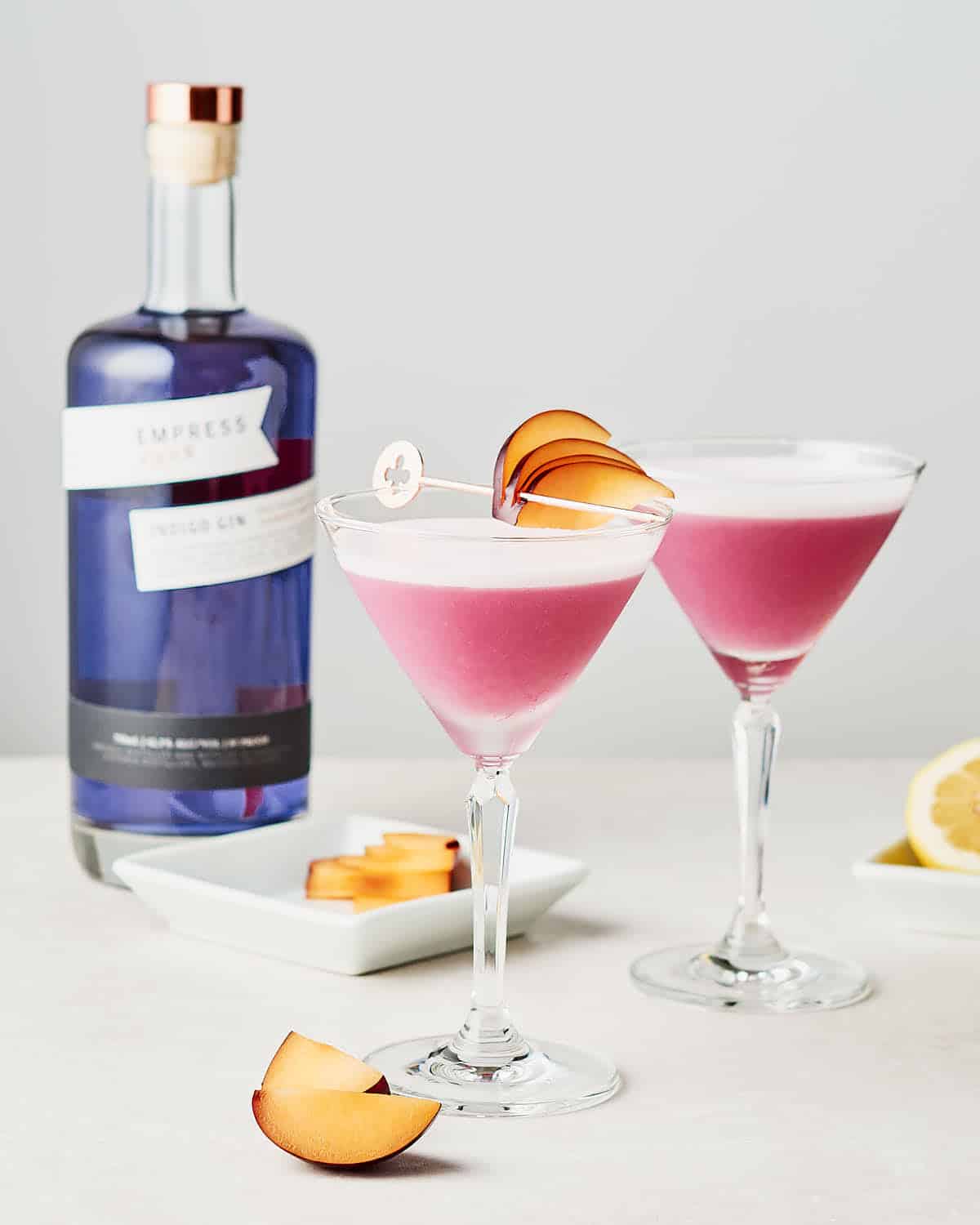 Side view of two cocktails of plum ginger gin sour with bottle of Empress Gin in background.
