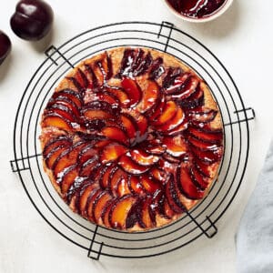 Overhead view of plum almond cake on black cake rack on grey surface with grey napkin and plums.