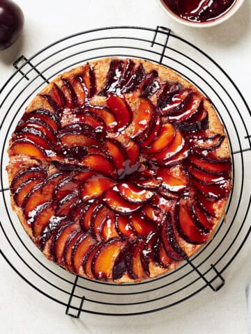 Overhead view of plum almond cake on black cake rack on grey surface with grey napkin and plums.