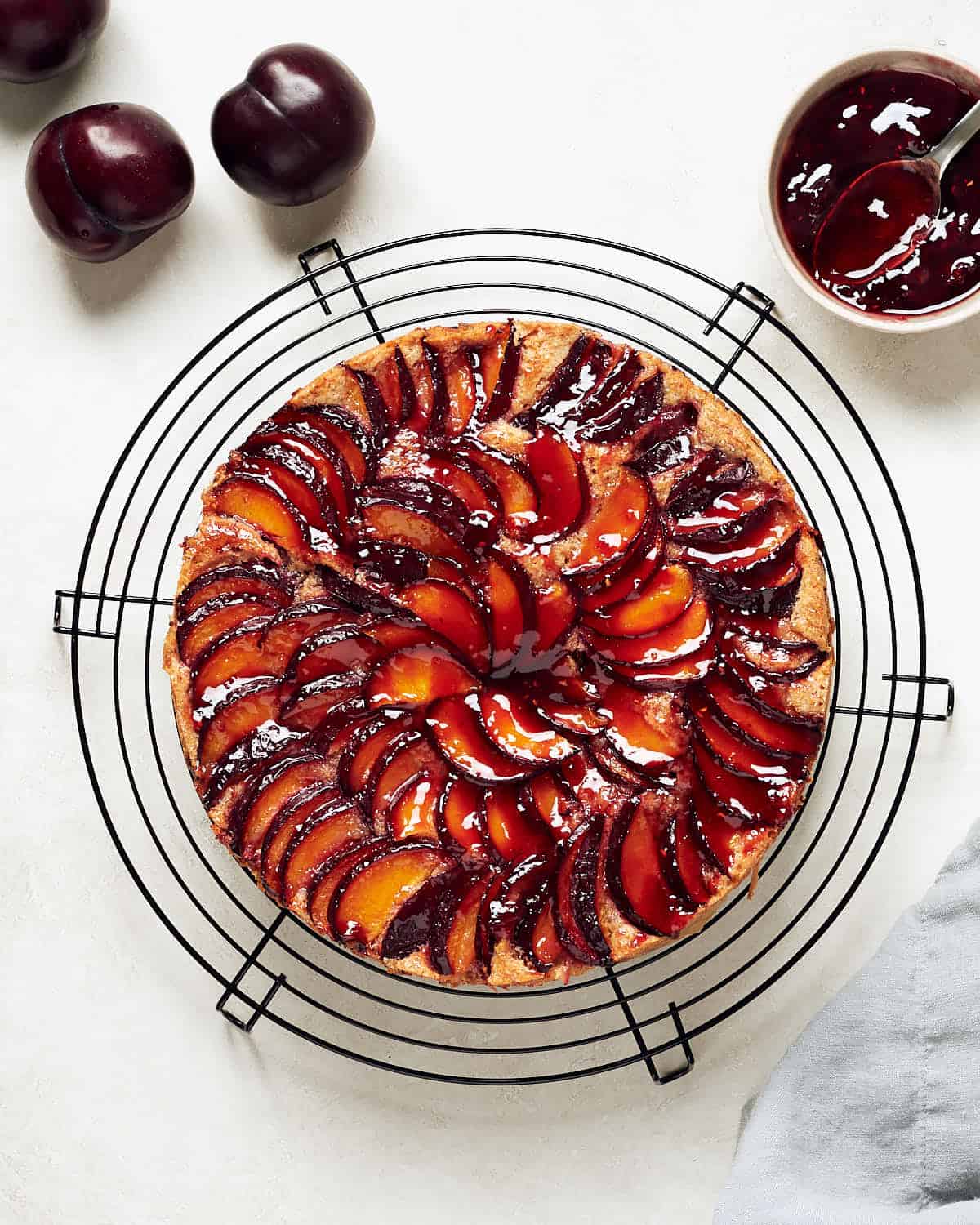 Overhead view of plum almond cake on black cake rack on grey surface with grey napkin, preserves, and plums.