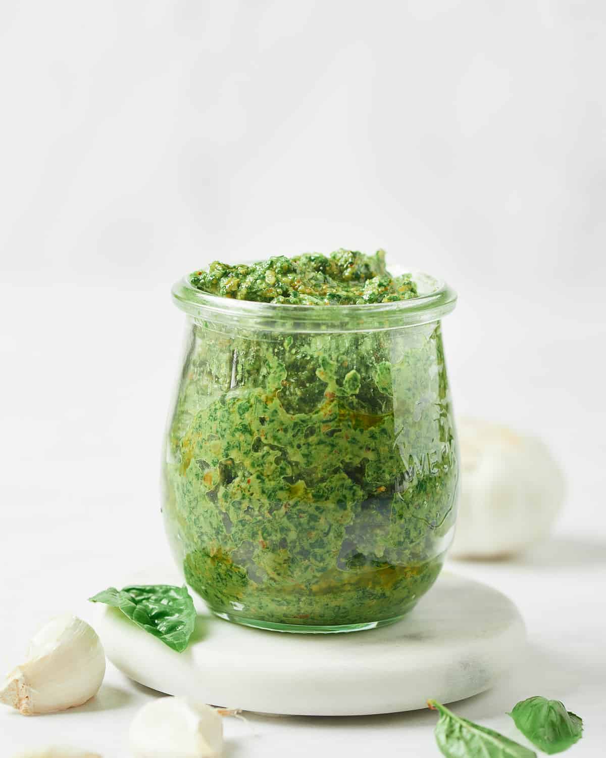 Side view of Vegan Walnut Pesto in glass jar with basil and garlic nearby.
