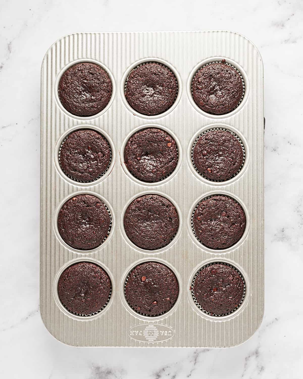 Overhead view of baked vegan chocolate fig cupcakes in cupcake liners in a muffin tin on a marble background.