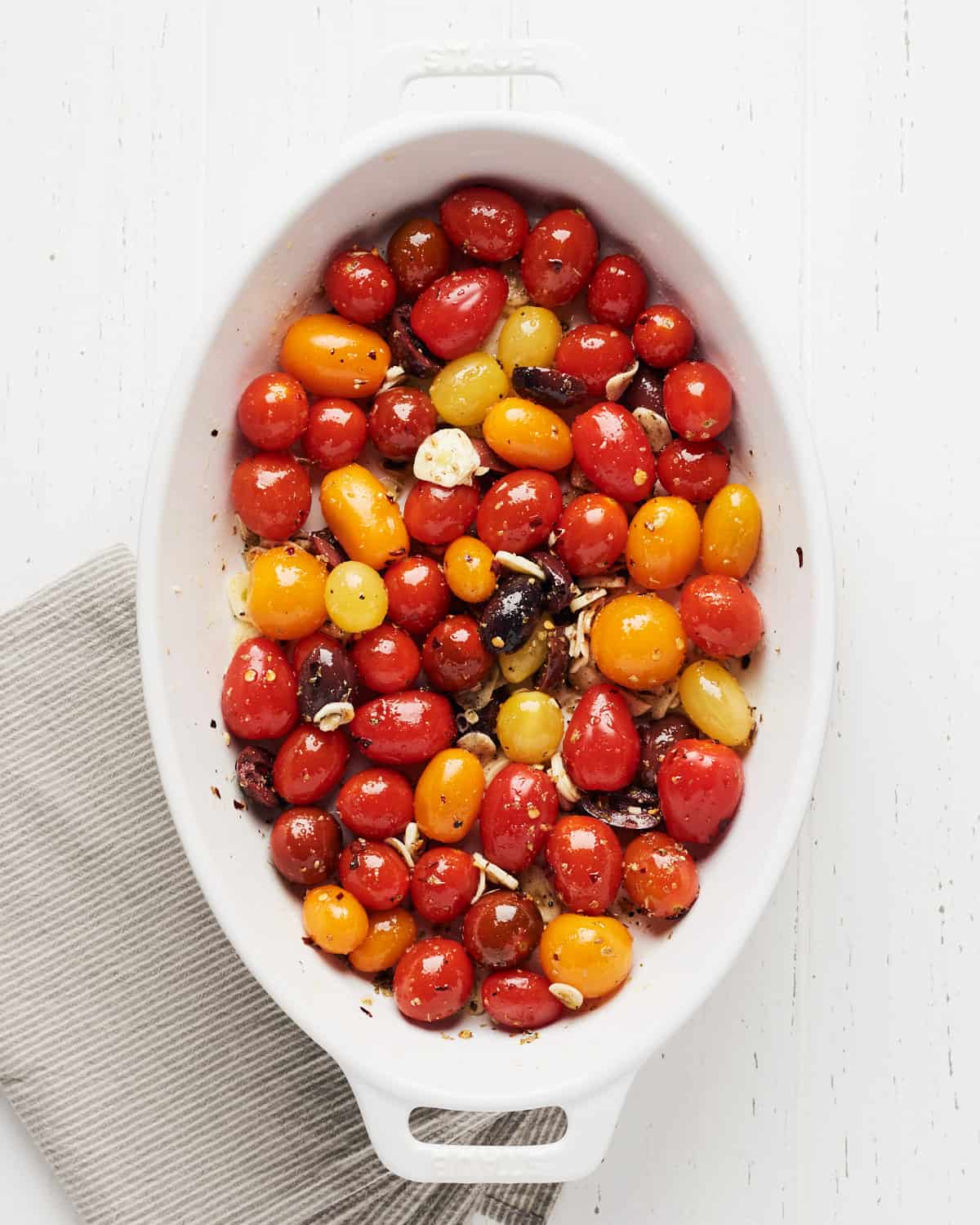Overhead view of fresh cherry tomatoes, sliced garlic, olives, spices, oil, and herbs mixed in a baking pan.