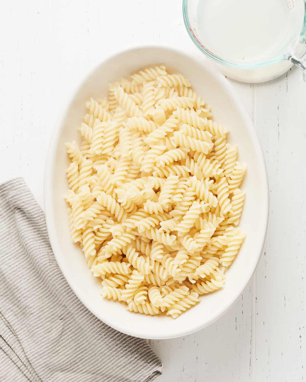 Overhead view of cooked fusilli in a white bowl with pasta water nearby on a white wood surface.