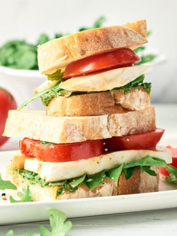 Side view of stacked Vegan Caprese Sandwich with sliced tomatoes, arugula, vegan pesto, and vegan mozzarella.