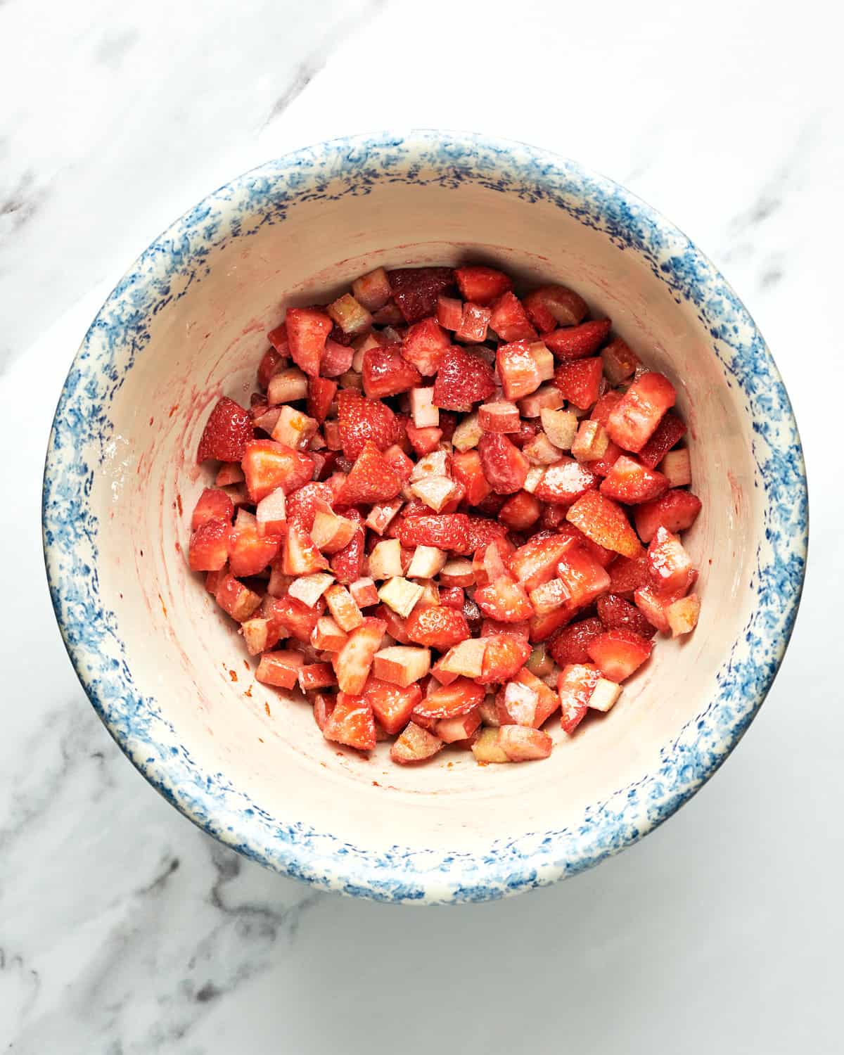 Overhead view of macerating strawberries and rhubarb in a bowl on a marble surface.