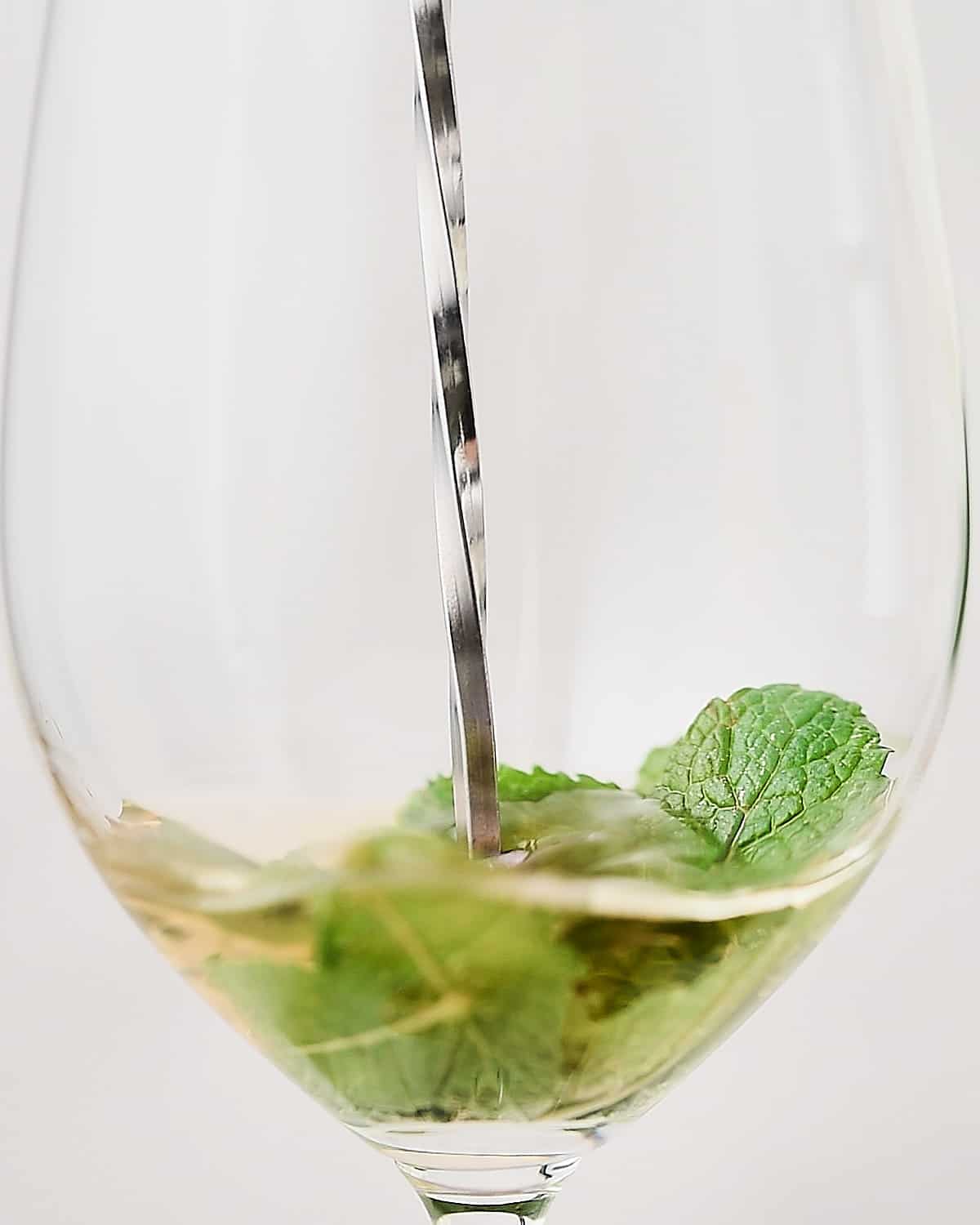 Muddling mint leaves with elderflower liqueur in a white wine glass.