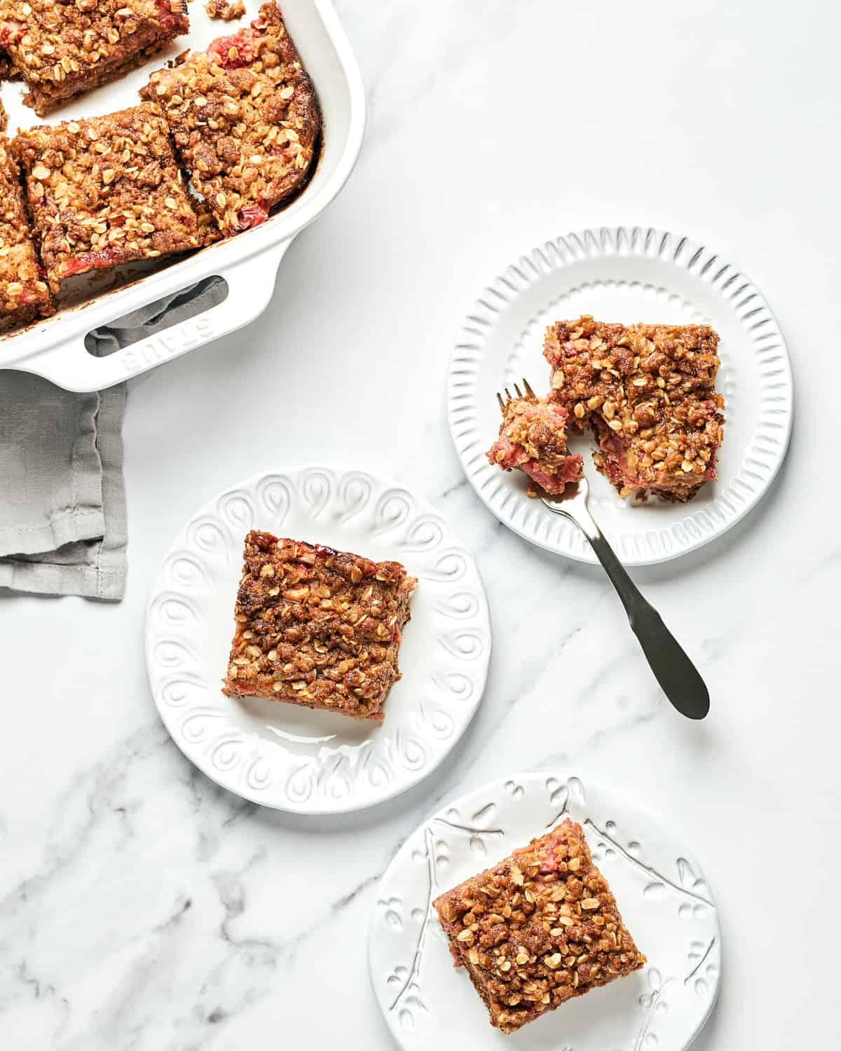 Overhead view of three plates of Vegan Strawberry Rhubarb Crumble Bars with a grey napkin and baking dish nearby.