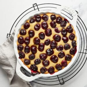 Overhead view of Vegan Clafoutis with Cherries in a white baking dish on a round black wire rack with a grey napkin.