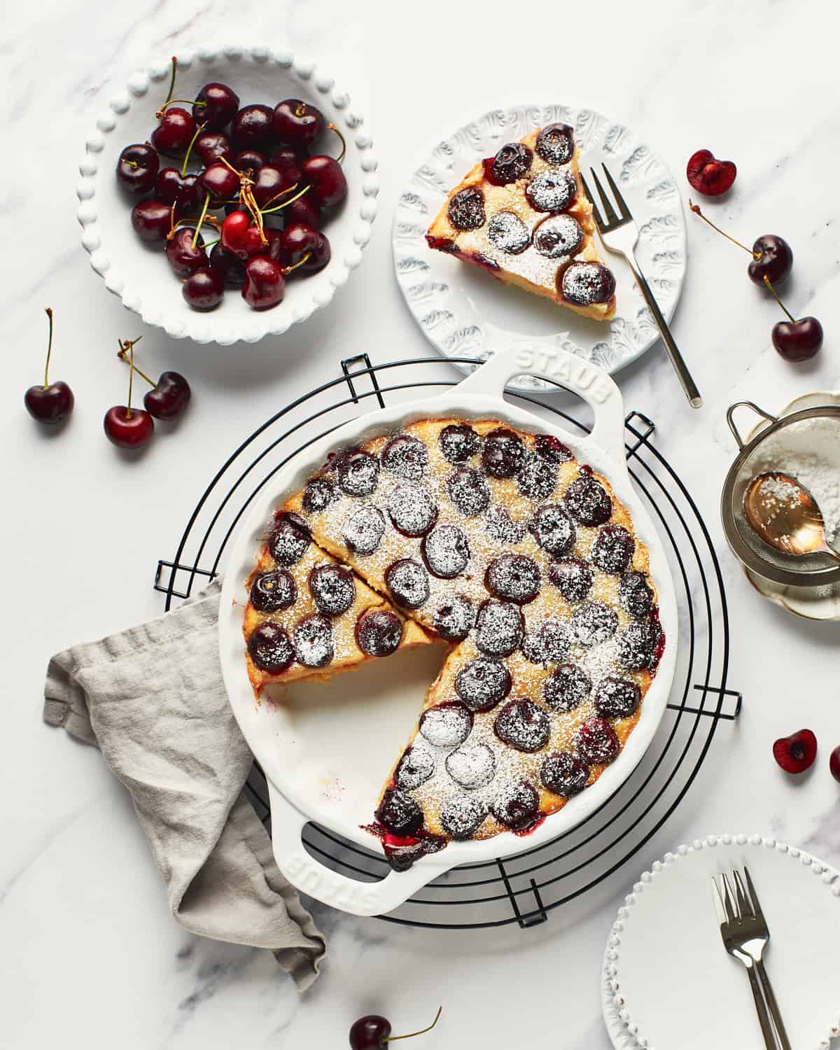 Overhead view of Vegan Clafoutis with a slice on a white plate with a small bowl of cherries.