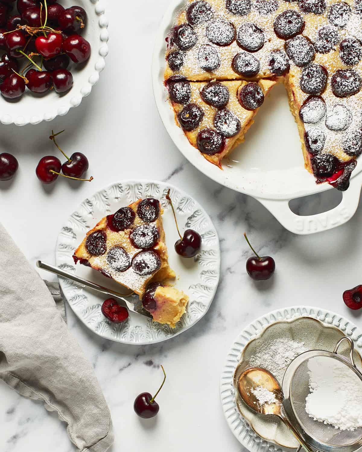Overhead view of vegan clafoutis slice on a plate with the rest in a pie plate with cherries on the side.
