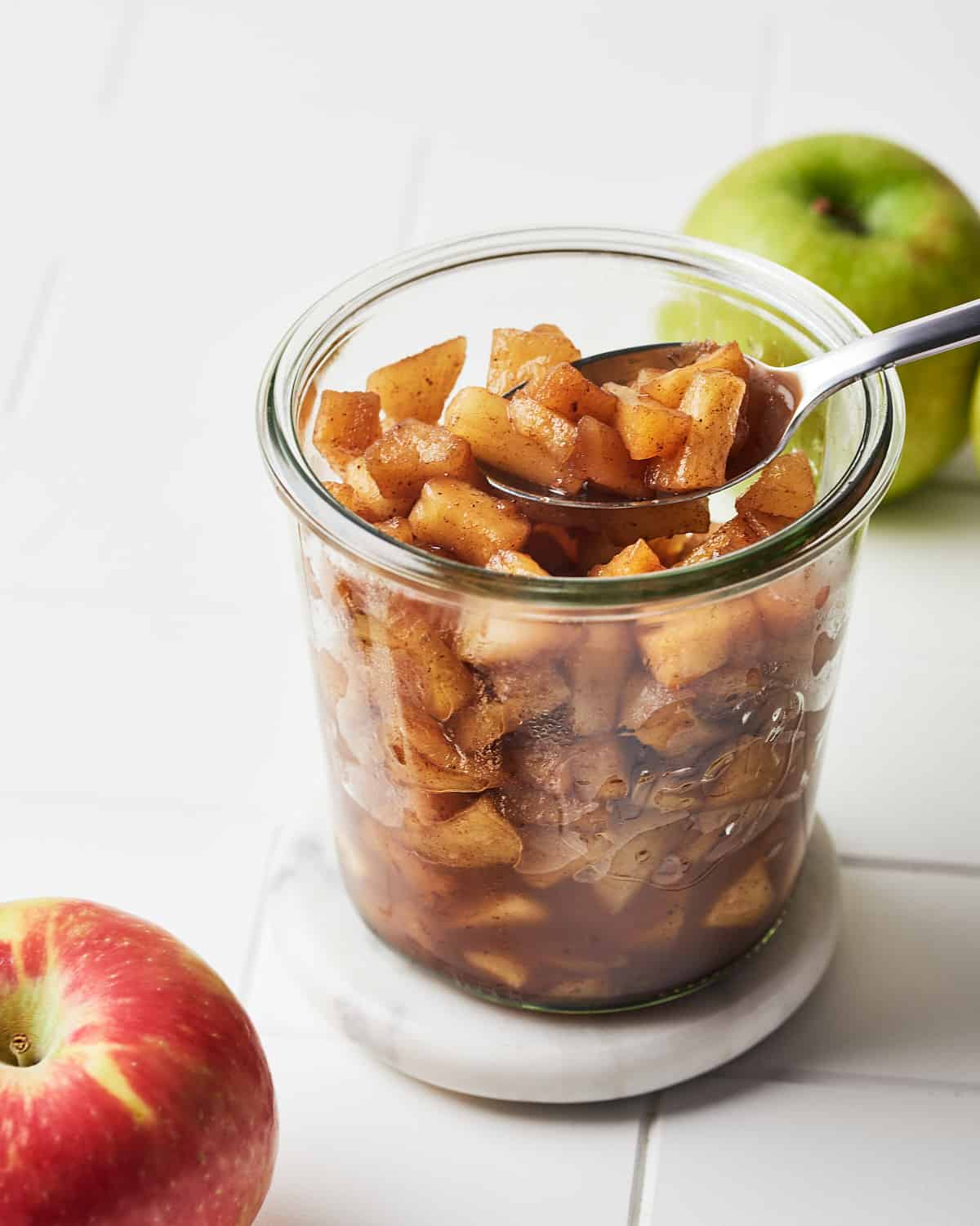 Side view of spoon with simple apple compote in a glass jar with 2 apples nearby.