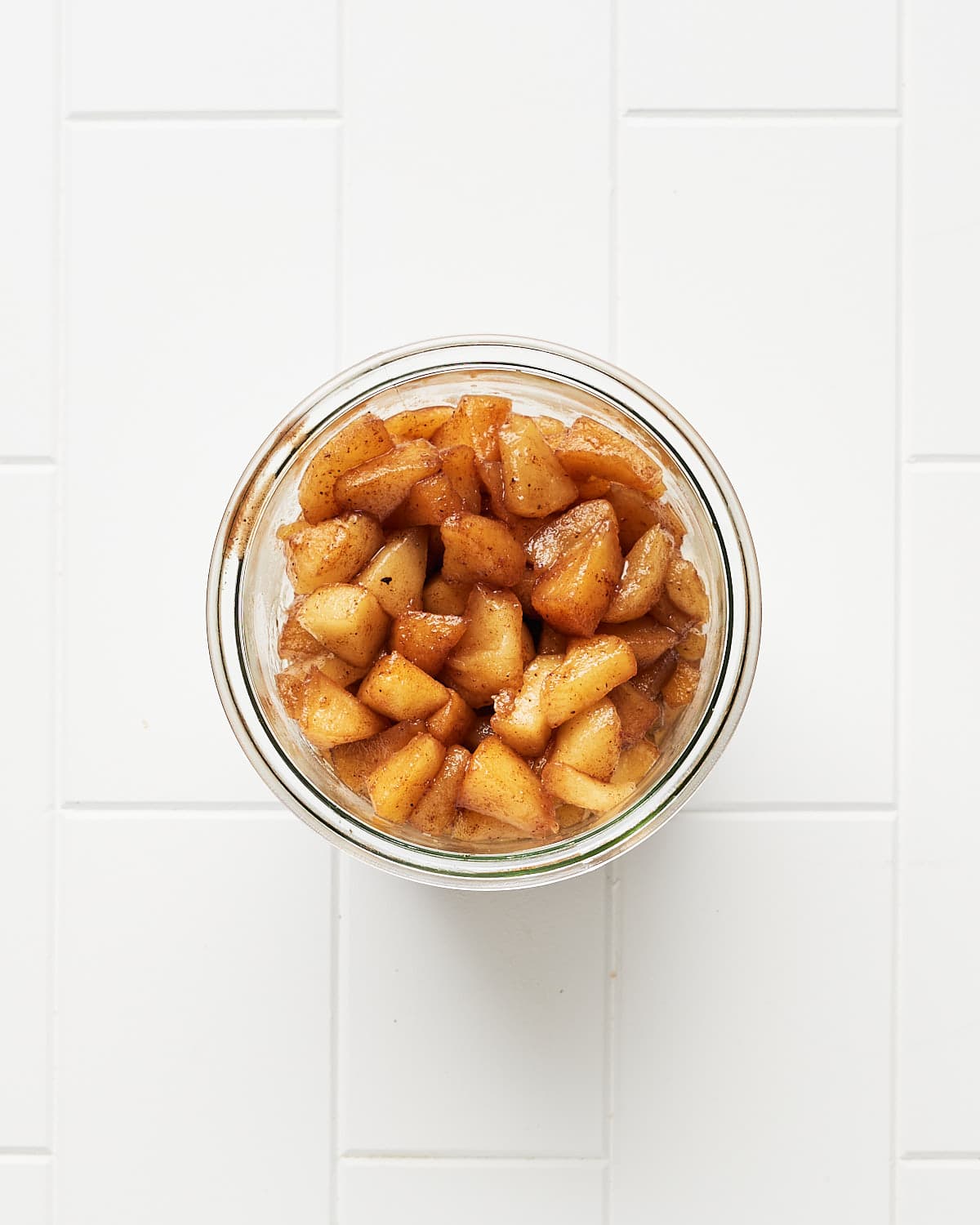 Overhead view of apple compote in a glass jar on a white surface.