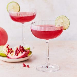 Side view of two glasses of pomegranate gimlets garnished with lime wheels.