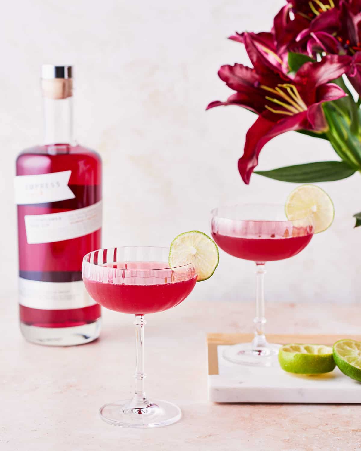 Side view of two glasses of pomegranate gin gimlet garnished with lime wheel next to a bottle of Empress 1908 Elderflower Rose Gin and red lilies.
