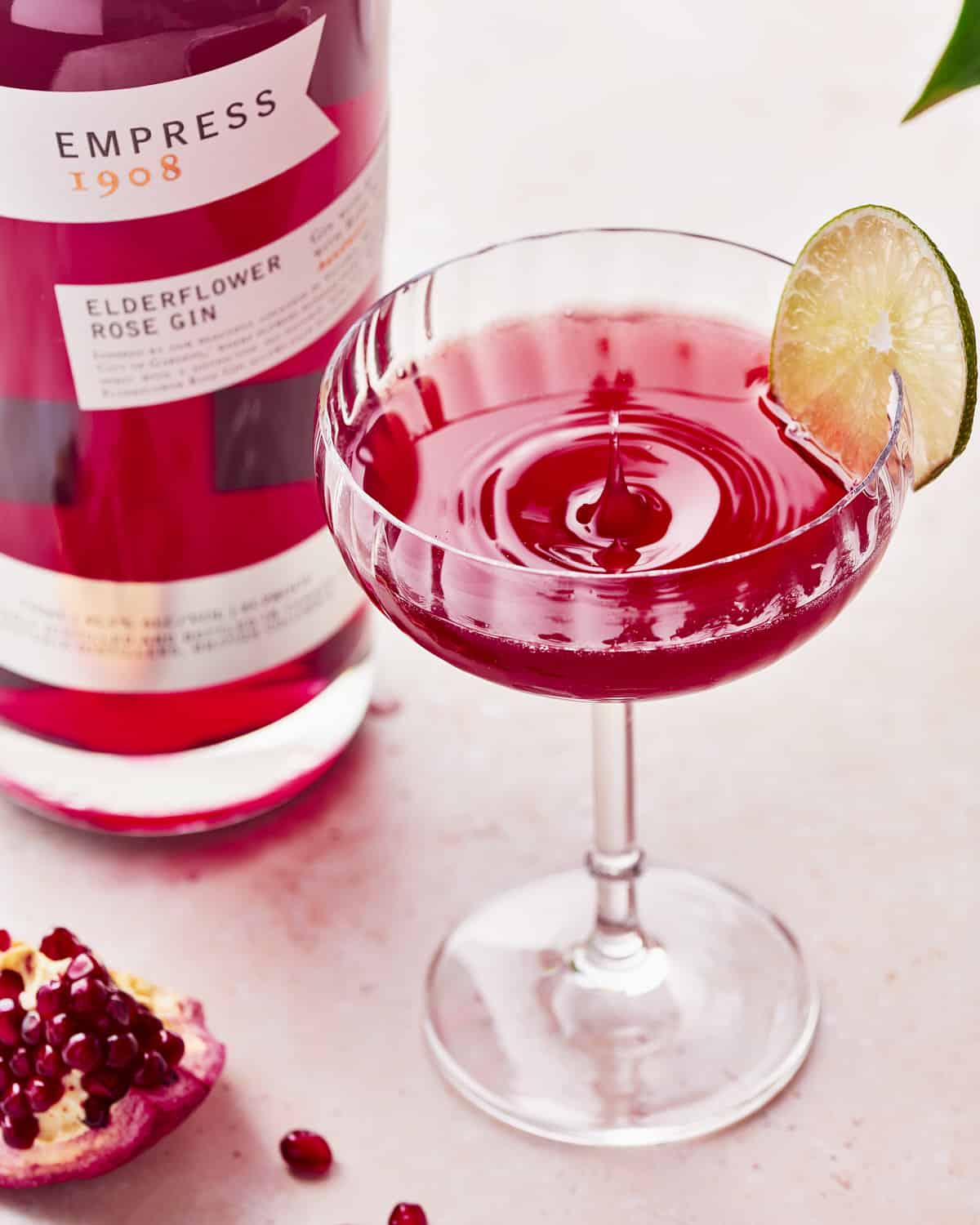 A splash of a glass of pomegranate gin gimlet garnished with lime wheel next to a bottle of Empress 1908 Elderflower Rose Gin.