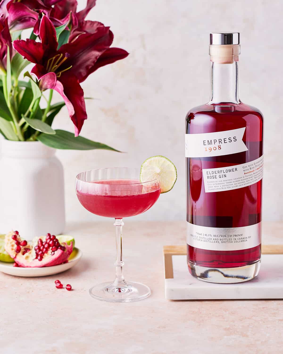 Side view of glass of pomegranate gin gimlet garnished with lime wheel next to a bottle of Empress 1908 Elderflower Rose Gin and red lilies.