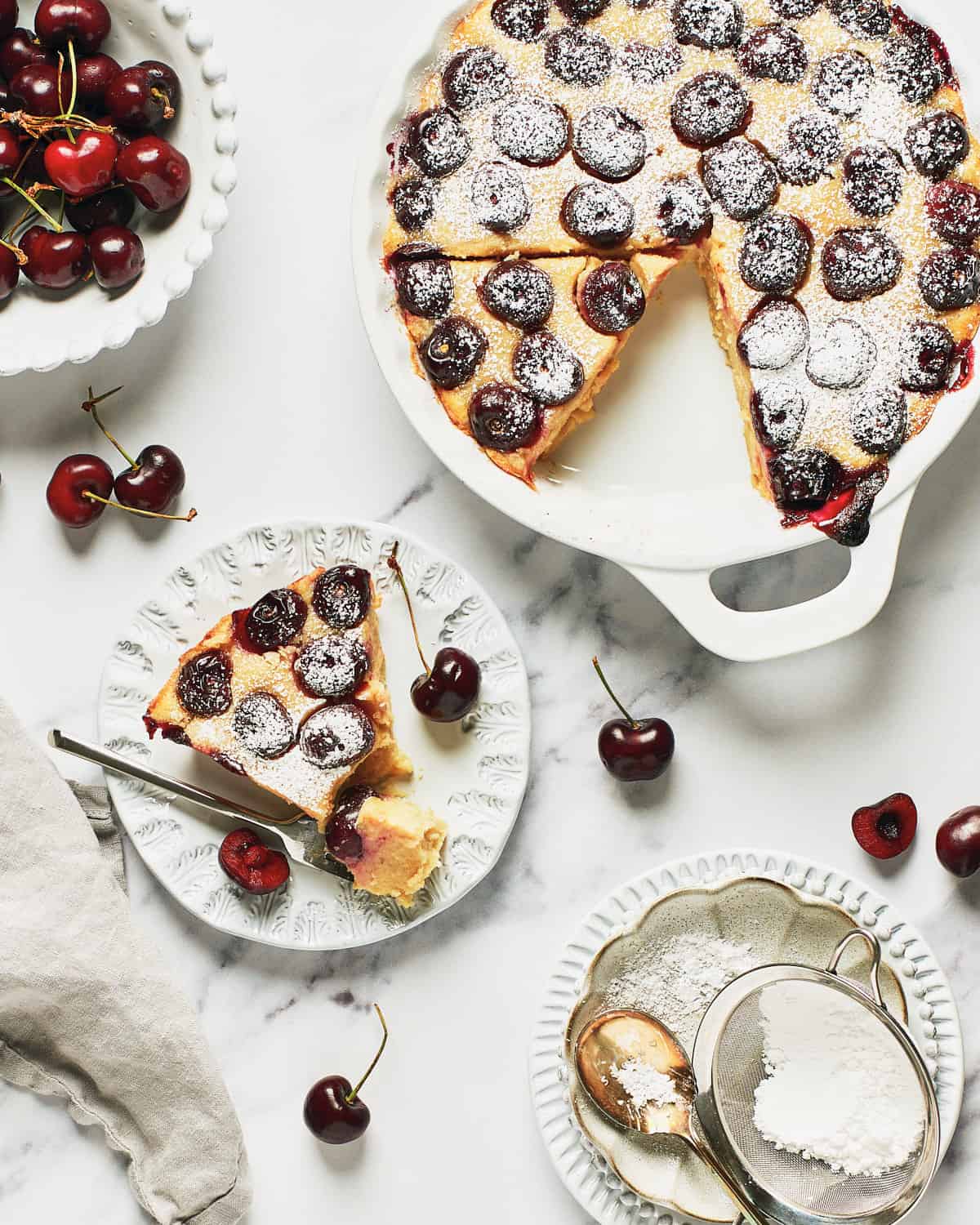 Overhead view of Vegan Clafoutis or Clafoutis Vegano with a slice on a white plate with a small bowl of cherries.