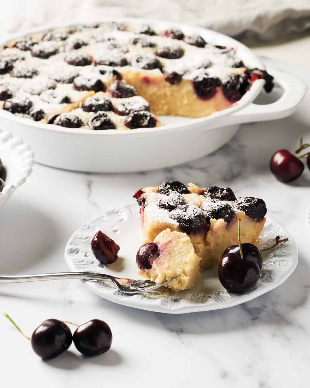 Slice of Vegan Clafoutis on a white plate with the rest of the clafoutis vegano in a pie plate next to it.