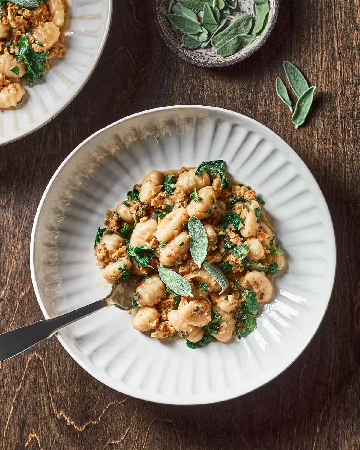Overhead view of gnocchi with vegan sausage, sage, and kale in a white bowl on a wooden surface with a fork.