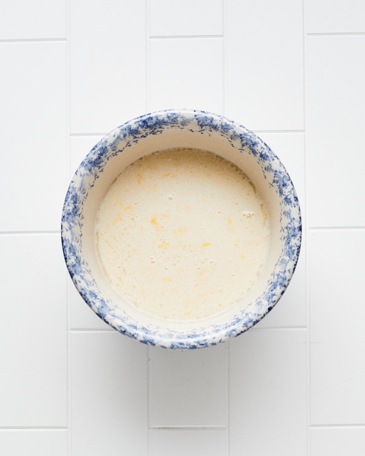 Overhead view of orange zest and liquid ingredients in a bowl on a while tile surface.