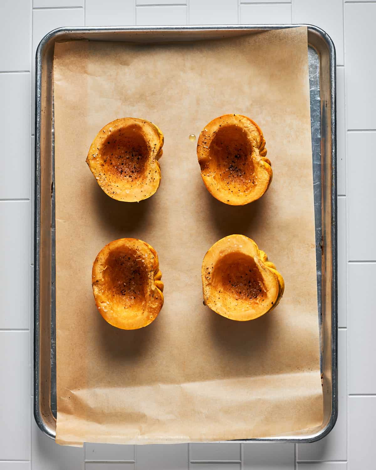 Overhead view of oiled carnival squash halves with salt and pepper on parchment paper on baking sheet before going in oven.