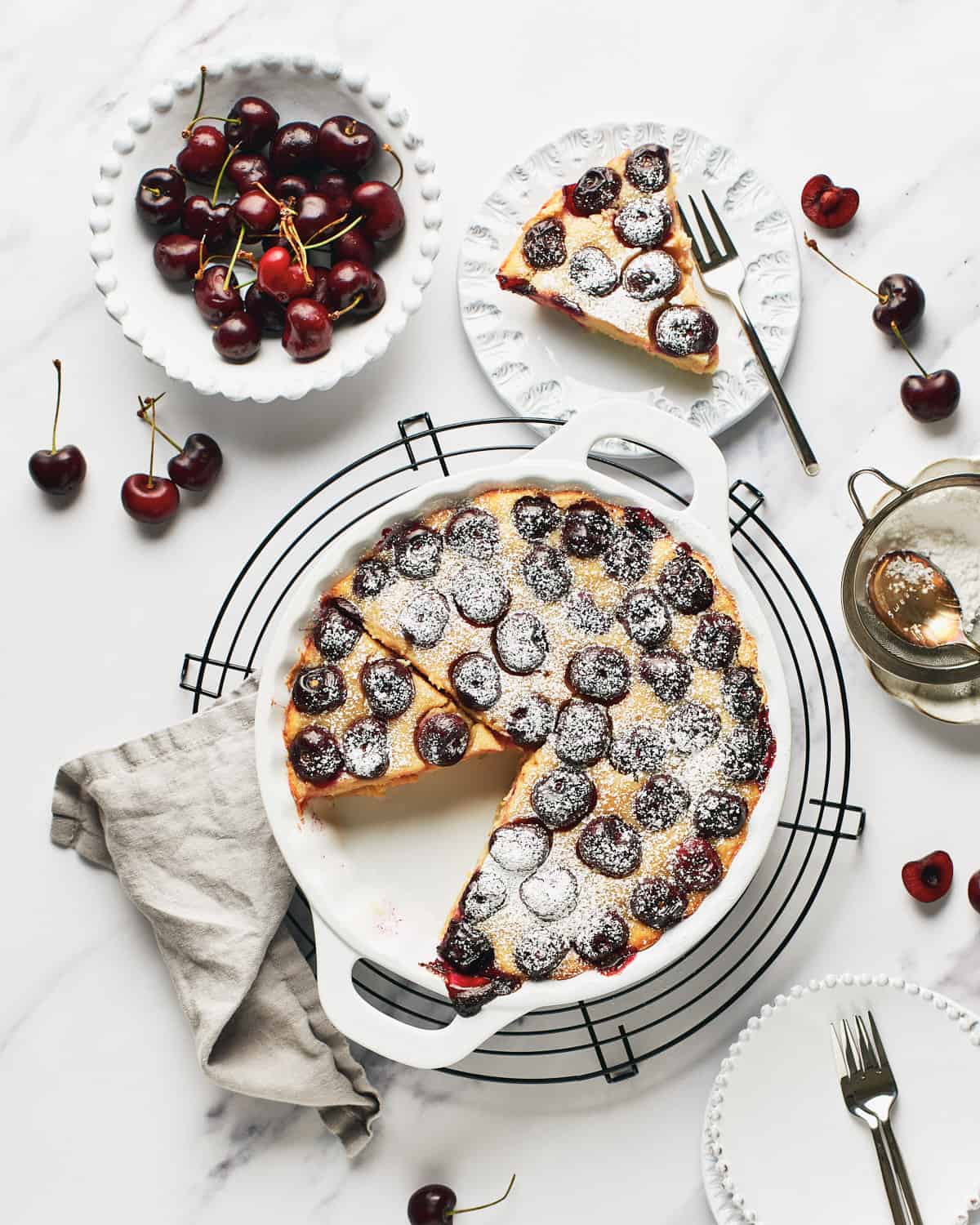 Overhead view of Vegan Clafoutis or Clafoutis Vegano with a slice on a white plate with a small bowl of cherries.