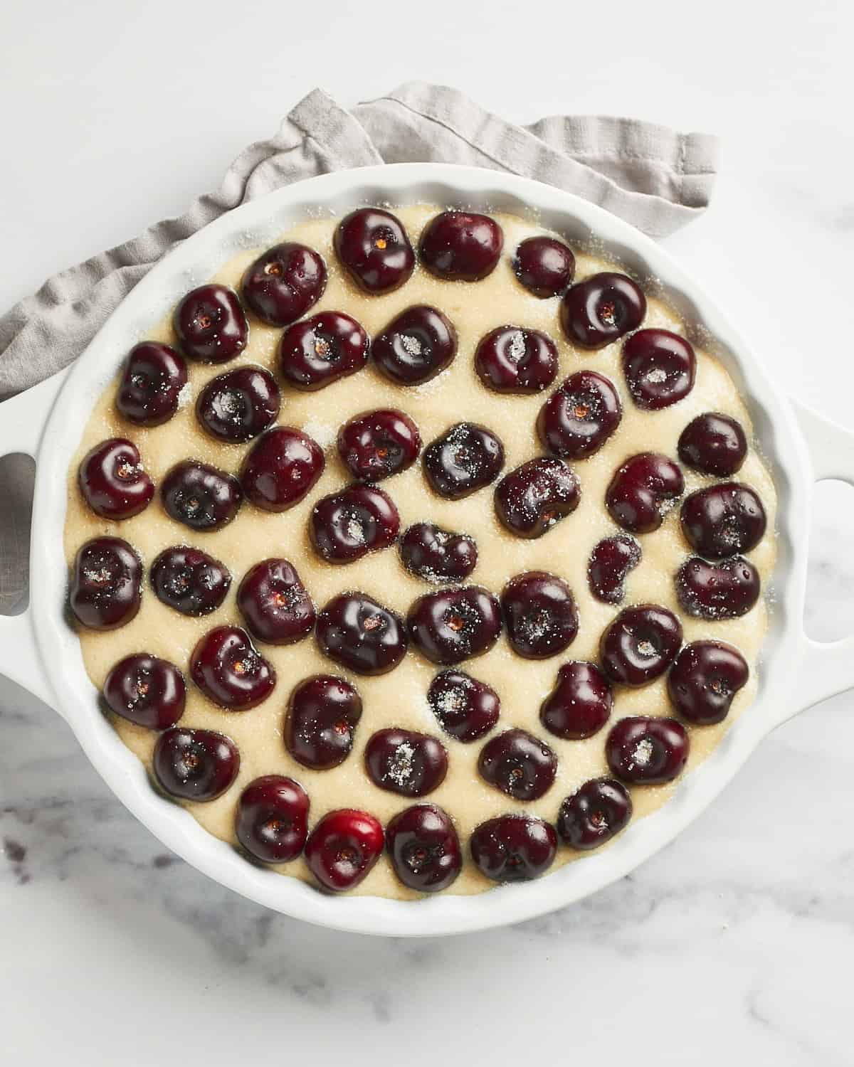 Overhead view of batter in a baking dish with fresh sweet cherries and sugar sprinkled on top.