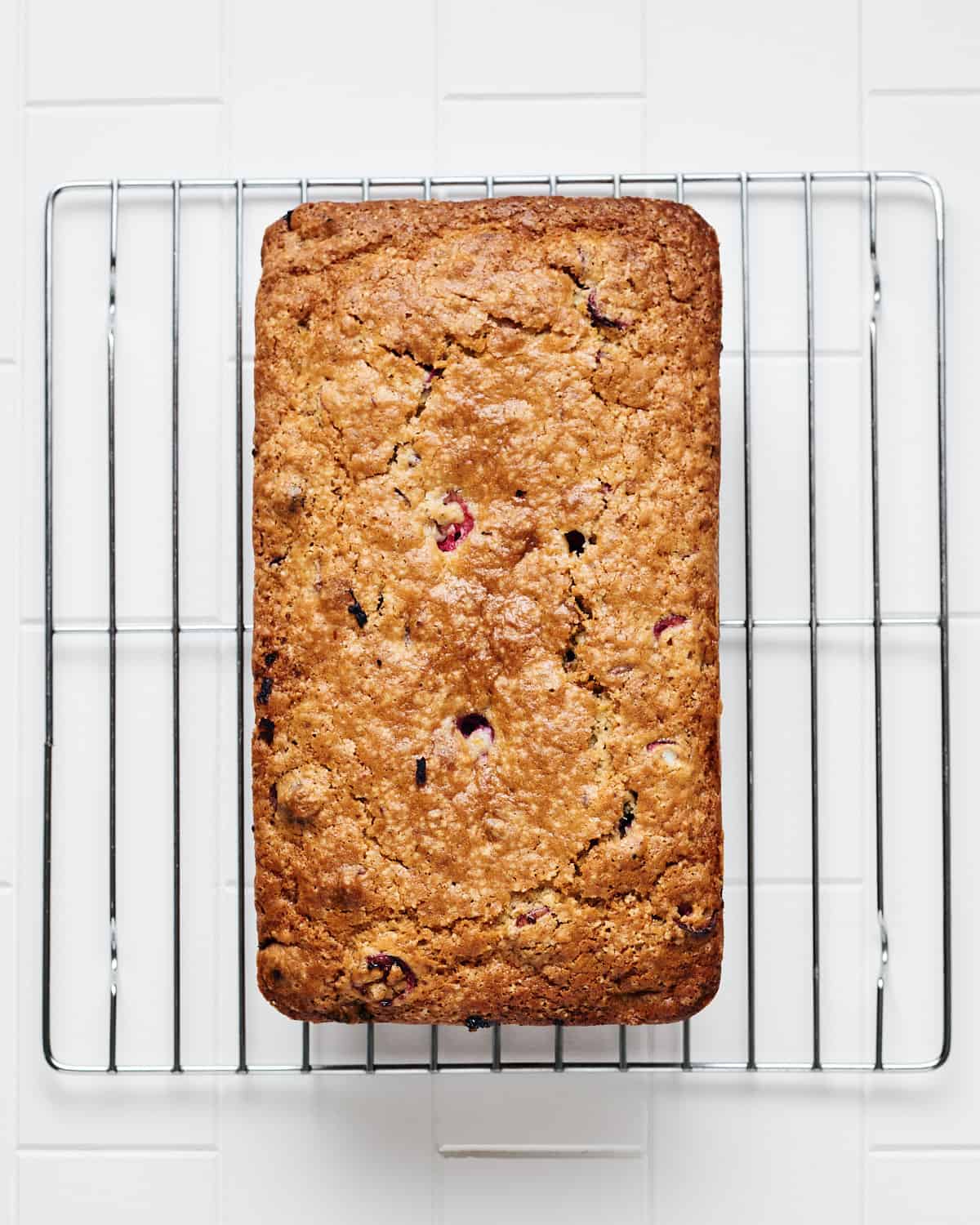 Overhead view of vegan cranberry orange bread cooling on a wire rack on a white tile surface.