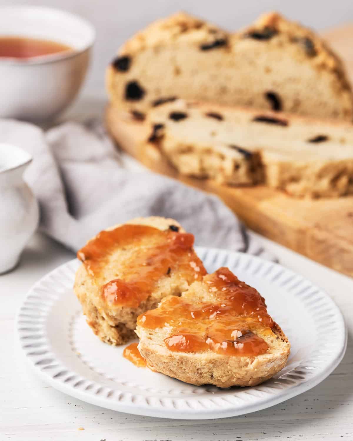 Slice of vegan Irish Soda Bread with vegan butter and jam with more slices on a cutting board with a cup of tea in the background.