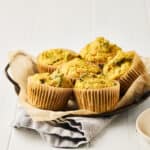Six vegan savory chickpea muffins with broccoli on parchment baking paper on a round metal tray with a grey napkin.