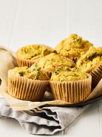 Six vegan savory chickpea muffins with broccoli on parchment baking paper on a round metal tray with a grey napkin.