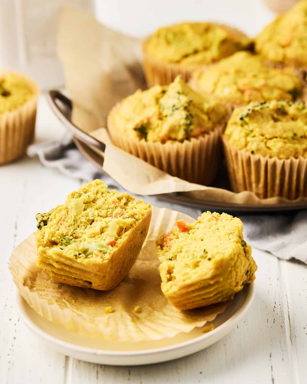 Vegan savory chickpea muffins with broccoli on parchment baking paper on a round metal tray behind one split muffin.