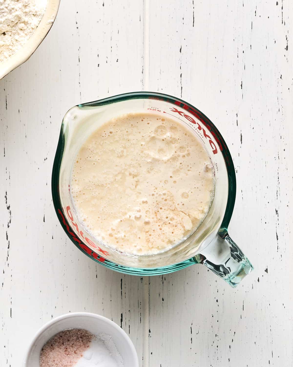 A measuring cup filled with soy milk and apple cider vinegar to make vegan buttermilk on a white wood surface with two bowls nearby.