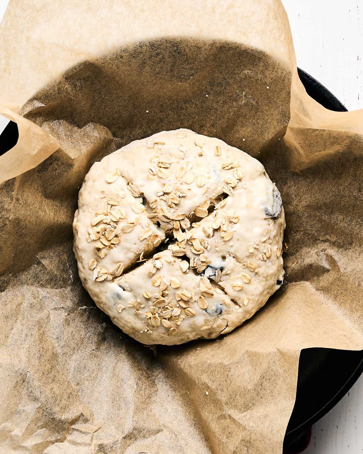 Unbaked dough with an X shaped on the top placed on parchment paper in a 9-inch cast iron pan on a white wooden surface.
