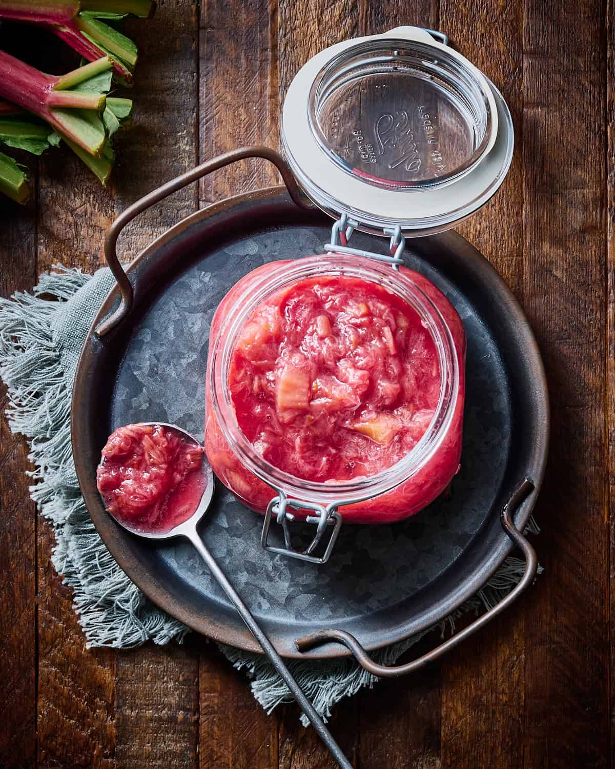 Rhubarb compote in a glass jar with spoonful next to it and stalks of rhubarb nearby.