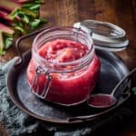 Rhubarb compote in a glass jar with spoonful next to it and stalks of rhubarb nearby.