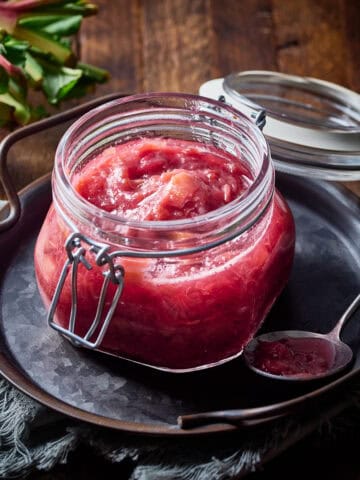 Rhubarb compote in a glass jar with spoonful next to it and stalks of rhubarb nearby.