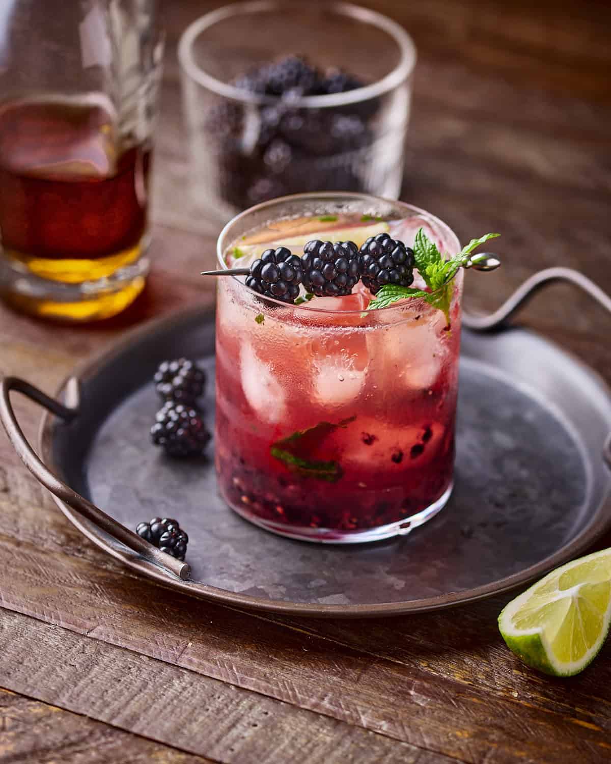 Glass of Blackberry Bourbon Smash garnished with blackberries and mint on a round serving plate on a wood surface.