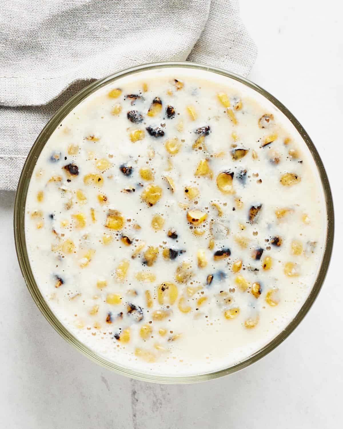 A mixture of liquid ingredients with corn in a glass bowl with a grey napkin on a marble surface.