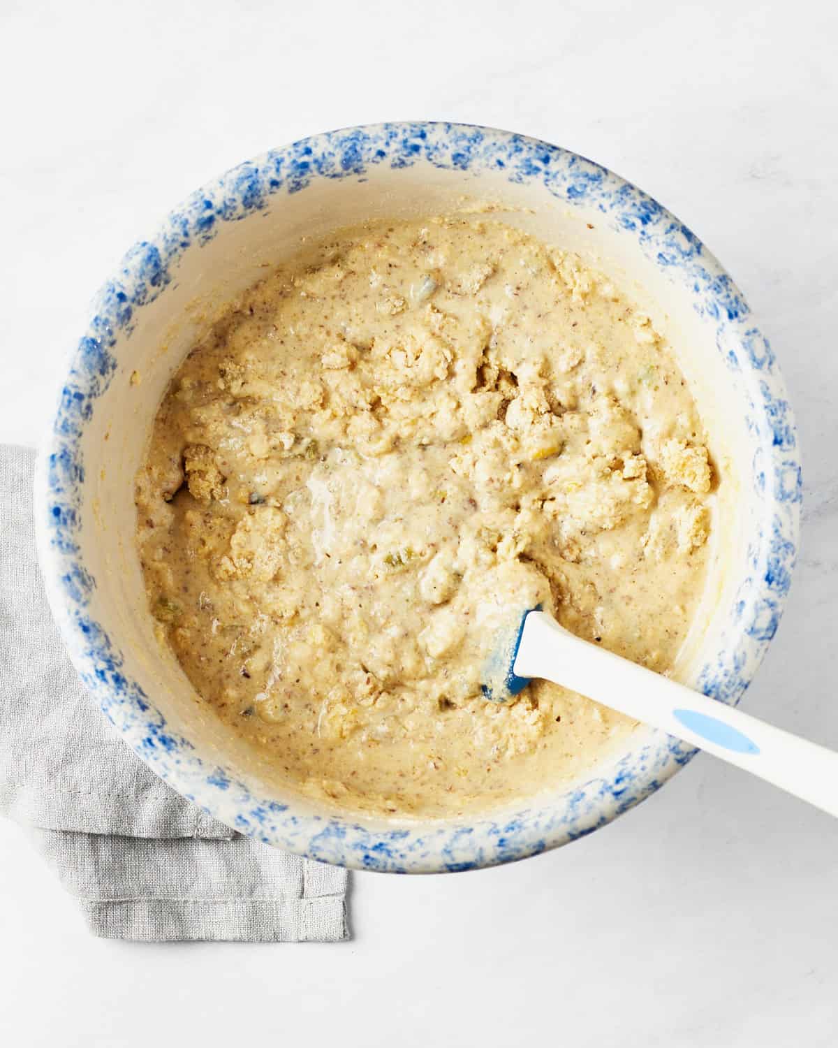 Liquid and dry ingredients mixed in a large bowl with a spatula on white marble background.